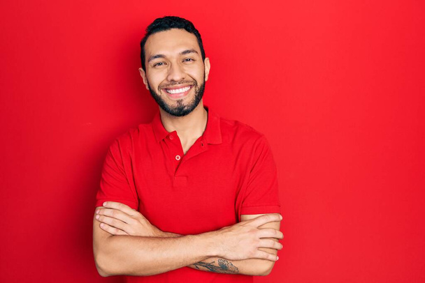 Hombre hispano con barba vistiendo casual camiseta roja cara feliz sonriendo con los brazos cruzados mirando a la cámara. persona positiva.  - Foto, Imagen