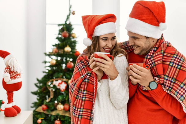 若いヒスパニック系のカップルはクリスマスの帽子をかぶって毛布で覆われています。自宅でコーヒーを飲みながら笑顔で立って. - 写真・画像
