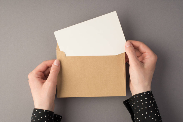 First person top view photo of female hands holding open craft paper envelope with white card on isolated grey background with blank space - Photo, Image
