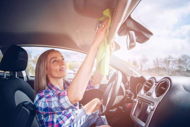 Mulher sorridente em camisa quadriculada limpa o interior do carro com um pano de microfibra. - Foto, Imagem