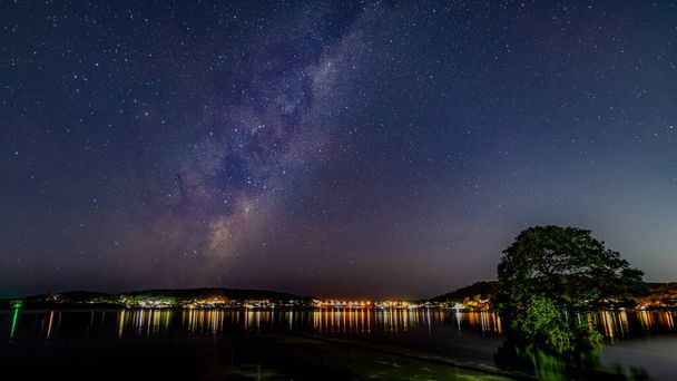Samanyolu ile gece gökyüzü, Avustralya, NSW 'nin merkez kıyısındaki Woy Woy rıhtımında ışık yansımaları ve ağaç. - Fotoğraf, Görsel