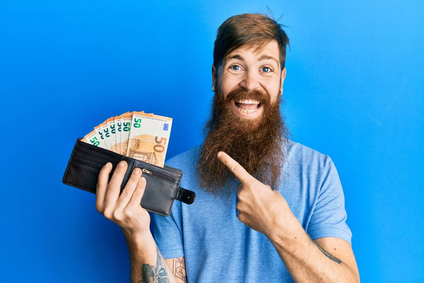 Hombre pelirrojo con barba larga sosteniendo billetera billetes en euros sonriendo feliz señalando con la mano y el dedo  - Foto, imagen