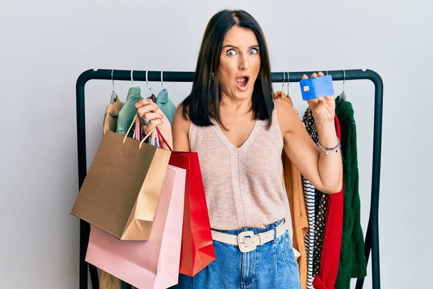 Middle age brunette personal shopper woman holding shopping bags and credit card afraid and shocked with surprise and amazed expression, fear and excited face.  - Photo, Image