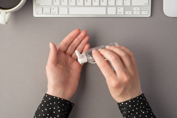First person top view photo of woman's hands using sanitizer transparent bottle white keyboard mouse and cup of coffee on isolated grey background - Foto, imagen