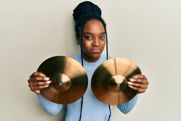 African american woman with braided hair holding golden cymbal plates skeptic and nervous, frowning upset because of problem. negative person.  - Photo, Image