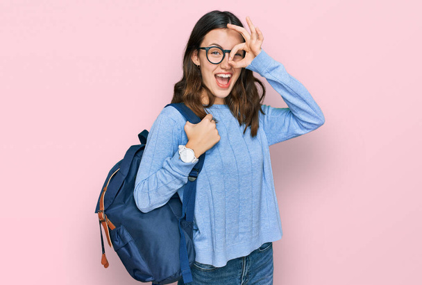 Νεαρό όμορφο κορίτσι φορώντας σακίδιο μαθητή χαμογελώντας χαρούμενος κάνει εντάξει σημάδι με το χέρι στο μάτι κοιτάζοντας μέσα από τα δάχτυλα  - Φωτογραφία, εικόνα