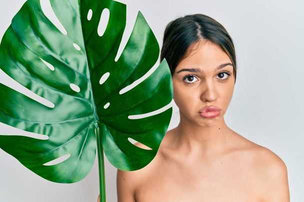 Young brunette woman holding green plant leaf close to beautiful face depressed and worry for distress, crying angry and afraid. sad expression.  - Fotó, kép