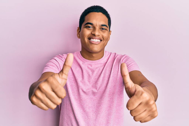 手で肯定的なジェスチャーを行うことを承認カジュアルなピンクのTシャツを着て若いハンサムなヒスパニック系の男性,親指を笑顔と成功のために幸せ.勝者のジェスチャー.  - 写真・画像