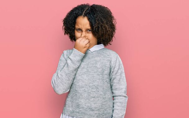 Young little girl with afro hair wearing casual clothes smelling something stinky and disgusting, intolerable smell, holding breath with fingers on nose. bad smell  - Photo, Image