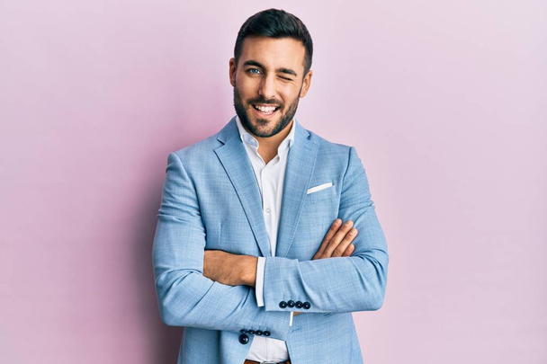 Junger hispanischer Geschäftsmann im Anzug mit verschränkten Armen zwinkert in die Kamera und blickt mit sexy Gesichtsausdruck, fröhlichem und glücklichem Gesicht.  - Foto, Bild