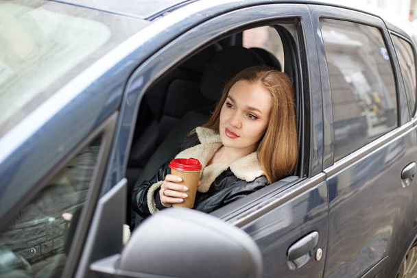 Una chica con el pelo largo está sentada en un coche negro, la vista desde la ventana. Tomando un vaso de café, pensando. - Foto, Imagen
