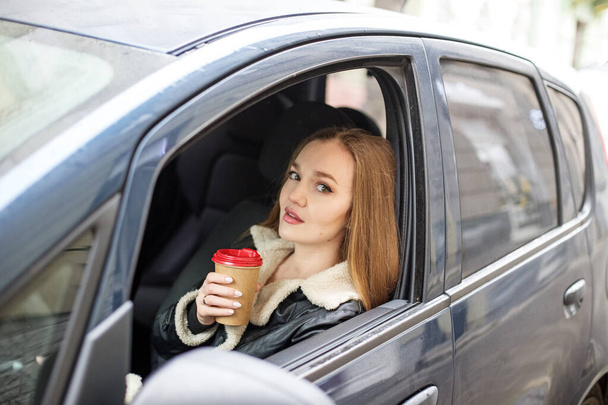 One girl with long hair is sitting in a black car, the view from the window. Holding a glass of coffee, thinking. - Foto, Imagen