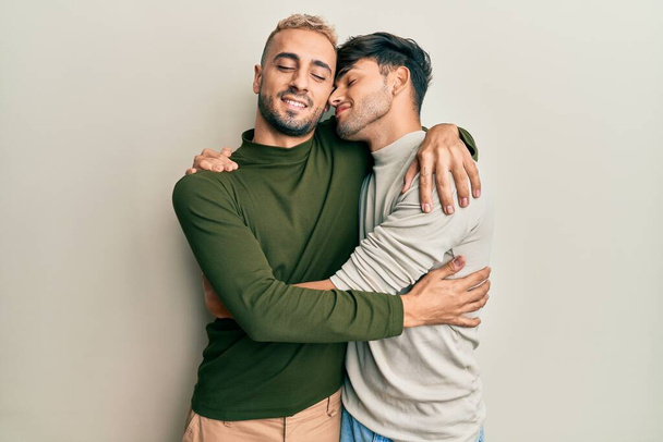 Ομοφυλόφιλο γκέι ζευγάρι που στέκεται μαζί φορώντας καθημερινά ρούχα αγκαλιάζοντας τον εαυτό του χαρούμενο και θετικό, χαμογελώντας με αυτοπεποίθηση. αυτοαγάπη και αυτοφροντίδα  - Φωτογραφία, εικόνα