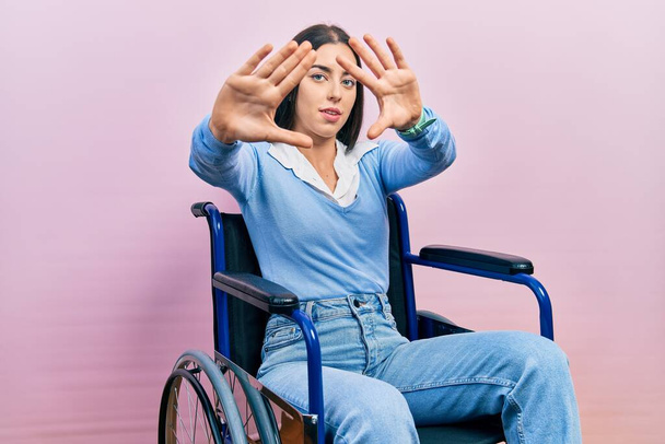 Mulher bonita com olhos azuis sentados em cadeira de rodas fazendo moldura usando as mãos palmas e dedos, perspectiva da câmera  - Foto, Imagem