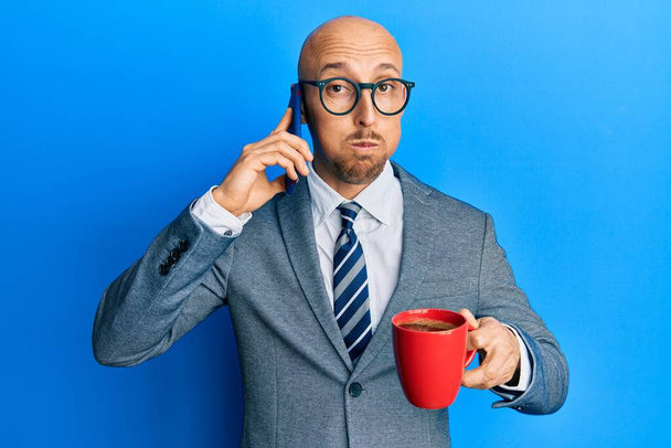 Homme chauve avec barbe à l'aide d'un smartphone et boire une tasse de café joues gonflées avec drôle de visage. bouche gonflée d'air, attrapant l'air.  - Photo, image