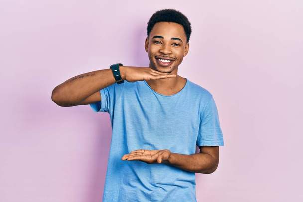 Молодой африканский американец в повседневной синей футболке жестикулирует руками, показывая большой и большой знак, измеряющий символ. улыбаясь, глядя в камеру. концепция измерения.  - Фото, изображение
