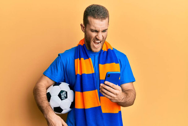 Молодой кавказский мужчина, держащий футбольный мяч, глядя на смартфон злой и безумный кричащий расстроенный и разъяренный, кричащий от гнева. гнев и агрессивная концепция.  - Фото, изображение
