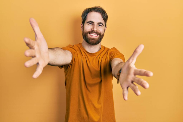 Homem caucasiano com barba vestindo camiseta amarela casual olhando para a câmera sorrindo de braços abertos para abraço. expressão alegre abraçando a felicidade.  - Foto, Imagem