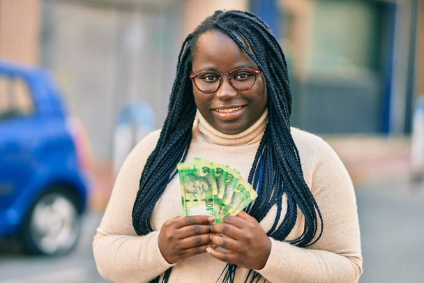 Joven mujer afroamericana sonriendo feliz sosteniendo billetes de rands sudafricanos en la ciudad. - Foto, imagen