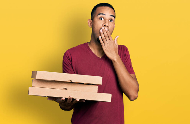 Junger afrikanisch-amerikanischer Mann, der Essen wegnimmt, das Mund und Hand bedeckt, geschockt und Angst vor Irrtümern. Überraschte Miene  - Foto, Bild