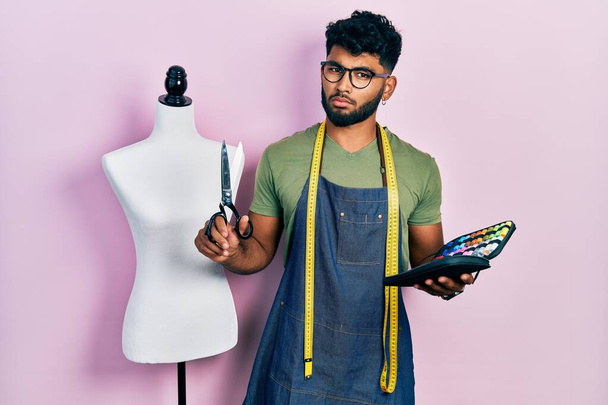 Arab man with beard dressmaker designer holding scissors and sewing kit clueless and confused expression. doubt concept.  - Photo, Image