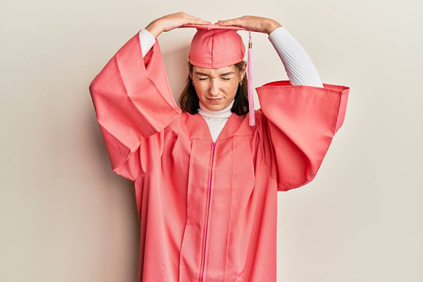 Young caucasian woman wearing graduation cap and ceremony robe suffering from headache desperate and stressed because pain and migraine. hands on head.  - Fotó, kép