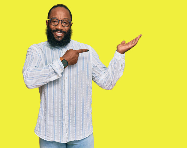 Młody Afroamerykanin w koszuli biznesowej i okularach zdumiony i uśmiechnięty do kamery, prezentujący się z ręką i wskazujący palcem.  - Zdjęcie, obraz