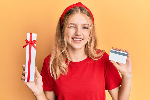 mooi jong Kaukasisch meisje holding geschenk en credit card knipogen naar de camera met sexy uitdrukking, vrolijk en gelukkig gezicht.  - Foto, afbeelding