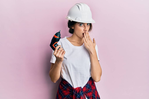 Nuori valkoihoinen nainen yllään arkkitehti kypärä käyttäen pora peittää suun käsin, järkyttynyt ja pelkää virhe. yllättynyt ilme  - Valokuva, kuva