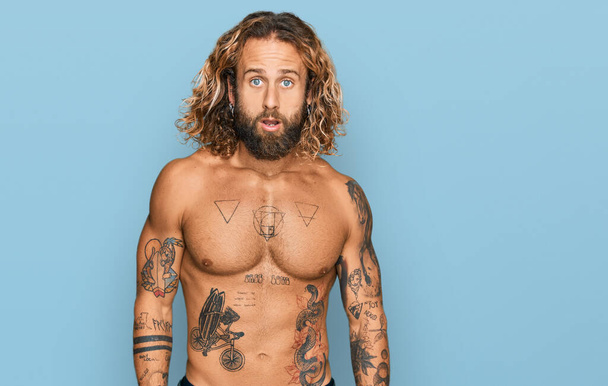 Handsome man with beard and long hair standing shirtless showing tattoos in shock face, looking skeptical and sarcastic, surprised with open mouth  - Photo, Image