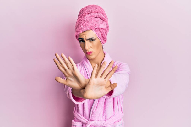 Young man wearing woman make up wearing shower towel on head and bathrobe rejection expression crossing arms and palms doing negative sign, angry face  - Photo, Image