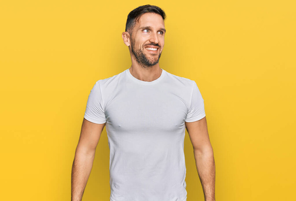 Handsome man with beard wearing casual white t shirt looking away to side with smile on face, natural expression. laughing confident.  - Photo, Image
