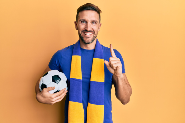 Jóképű férfi szakállas futball huligán éljenzés játék gazdaság labda mosolyogva egy ötlet vagy kérdés ujjal mutogató boldog arc, első számú  - Fotó, kép