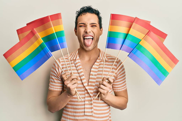 Schöner Mann mit Make-up, der Regenbogen-Lgbtq-Flaggen hochhält, die die Zunge mit lustigem Gesichtsausdruck herausstrecken.  - Foto, Bild