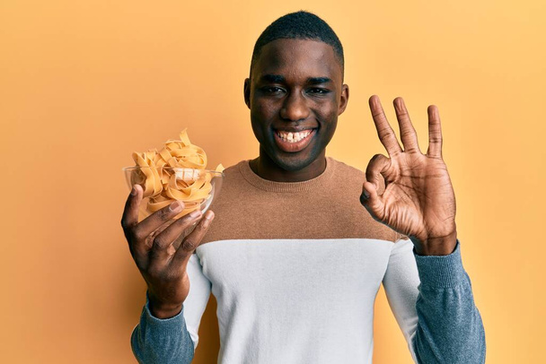 Молодой африканский американец держит миску с сырыми спагетти делает хорошо знак с пальцами, улыбаясь дружелюбный жест отличный символ  - Фото, изображение