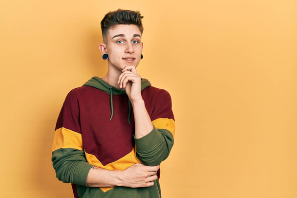 Young caucasian boy with ears dilation wearing casual sweatshirt looking confident at the camera with smile with crossed arms and hand raised on chin. thinking positive.  - Photo, Image