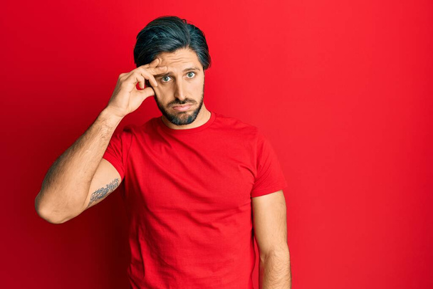 Joven hombre hispano con camiseta roja casual preocupado y estresado por un problema con la mano en la frente, nervioso y ansioso por la crisis  - Foto, imagen