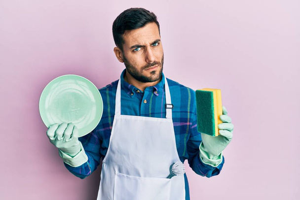 Jonge Spaanse man draagt schort met geurmaker die de afwas doet sceptisch en nerveus, fronsend van streek vanwege het probleem. negatieve persoon.  - Foto, afbeelding