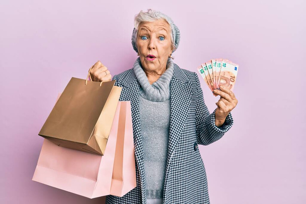 Oudere grijsharige vrouw die winkeltassen en eurobankbiljetten in shock houdt en er sceptisch en sarcastisch uitziet, verrast met open mond  - Foto, afbeelding