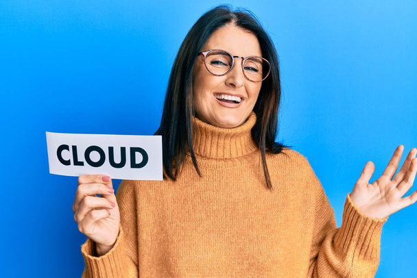 Middle age brunette woman holding paper with cloud word celebrating achievement with happy smile and winner expression with raised hand  - Fotoğraf, Görsel