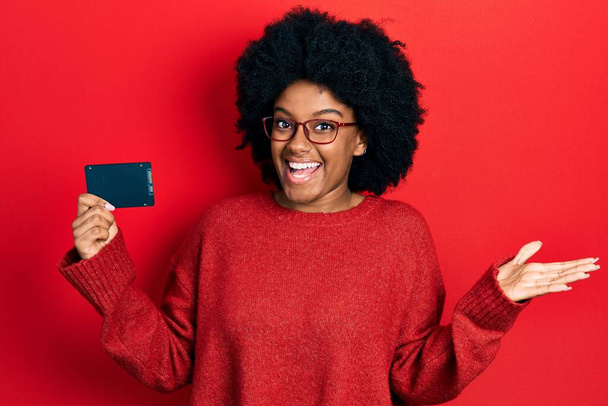 Young african american woman holding ssd memory celebrating achievement with happy smile and winner expression with raised hand  - Фото, изображение