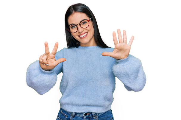 Giovane donna ispanica indossa abiti casual e occhiali che mostrano e puntano verso l'alto con le dita numero otto sorridendo fiducioso e felice.  - Foto, immagini
