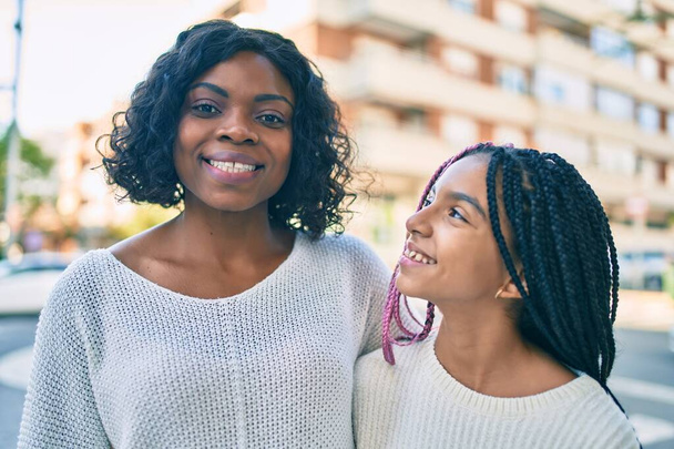 Όμορφη Αφροαμερικανή μητέρα και κόρη χαμογελάνε χαρούμενες και αγκαλιάζονται. Στέκεται με χαμόγελο στο πρόσωπο στέκεται στην πόλη. - Φωτογραφία, εικόνα
