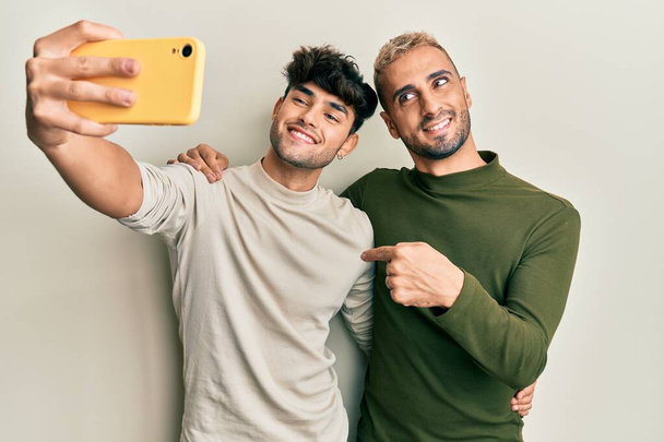 Homoseksüel eşcinsel çift bir arada durup akıllı telefonlu bir fotoğraf çekiyorlar mutlu ve gururlu bir şekilde gülümseyen birini işaret ediyorlar.  - Fotoğraf, Görsel