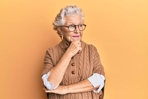 Starší šedovlasá žena v neformálním oblečení a brýlích s rukou na bradě přemýšlející o otázce a zamyšleném výrazu. s zamyšleným úsměvem. pochybný koncept.  - Fotografie, Obrázek