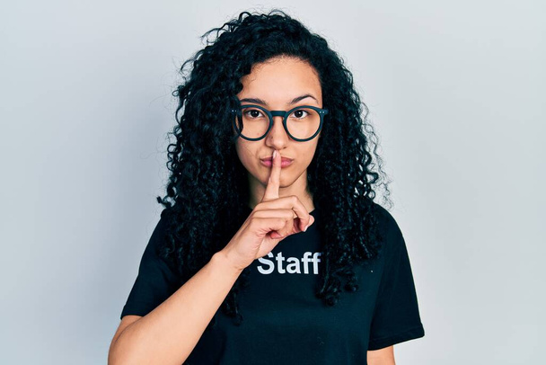 Jonge Latijns-Amerikaanse vrouw met krullend haar draagt personeel t shirt vragen om stil te zijn met de vinger op de lippen. stilte en geheim concept.  - Foto, afbeelding