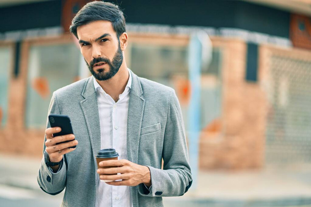 Νεαρός Ισπανός επιχειρηματίας με σοβαρή έκφραση χρησιμοποιώντας smartphone και πίνοντας καφέ στην πόλη. - Φωτογραφία, εικόνα