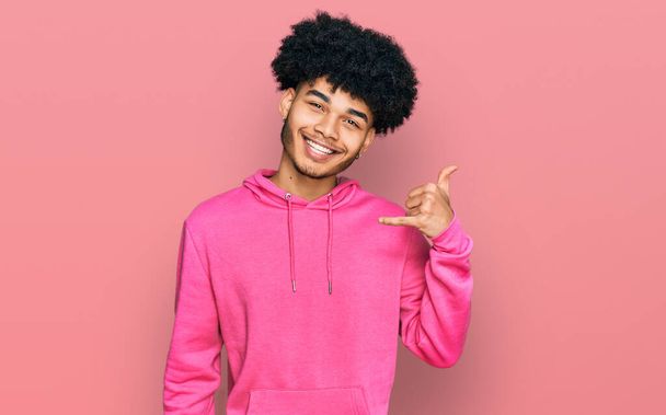 Young african american man with afro hair wearing casual pink sweatshirt smiling doing phone gesture with hand and fingers like talking on the telephone. communicating concepts.  - Photo, Image