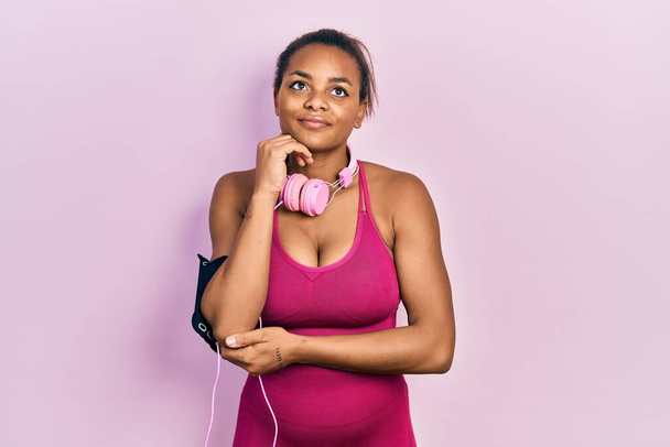 Young african american girl wearing gym clothes and using headphones with hand on chin thinking about question, pensive expression. smiling and thoughtful face. doubt concept.  - Photo, Image