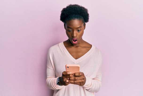 Молодая африканская американская девушка, используя смартфон боится и шокирован удивлением и удивлением выражение лица, страх и возбужденное лицо.  - Фото, изображение